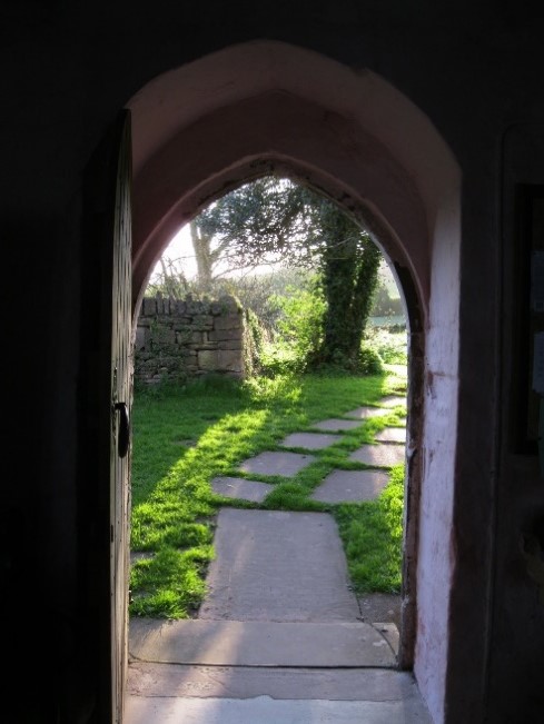 A photograph of an open door, the door opening inwards. The view outside is of a garden or yard, thick with green grass and paving stones laid out to form a path off and to the right. There is an old stone wall just beyond the doorway and to the left, next to a tree covered in ivy.