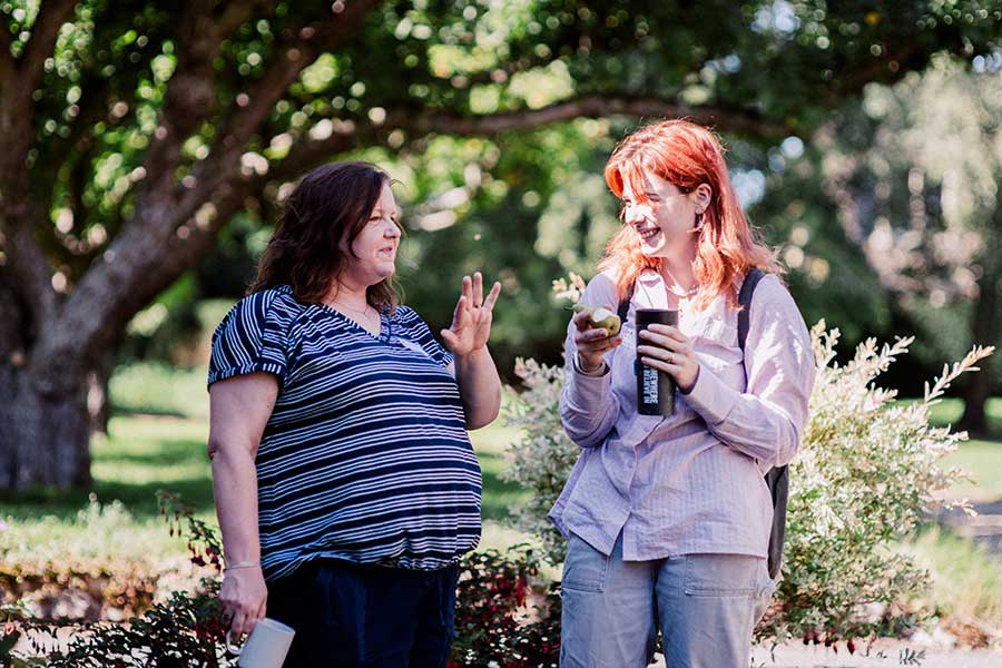 Two women chatting in the sunshine. They are both caucasian. One has red hair and the other had dark brown hair.