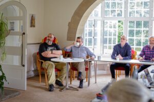Four men sitting in a room on chairs and leaning on tables. They are listening to a lecture or presentation.