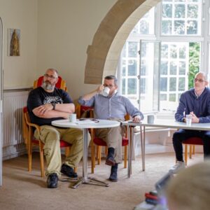 Four men sitting in a room on chairs and leaning on tables. They are listening to a lecture or presentation.
