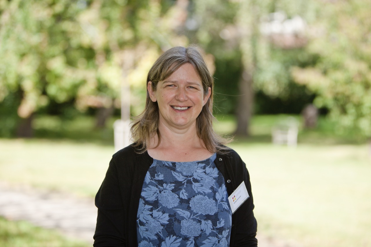 Jenny Corcoran standing outside on grass. Jenny has shoulder length brown hair and is wearing a black cardigan over a blue flowery dress.