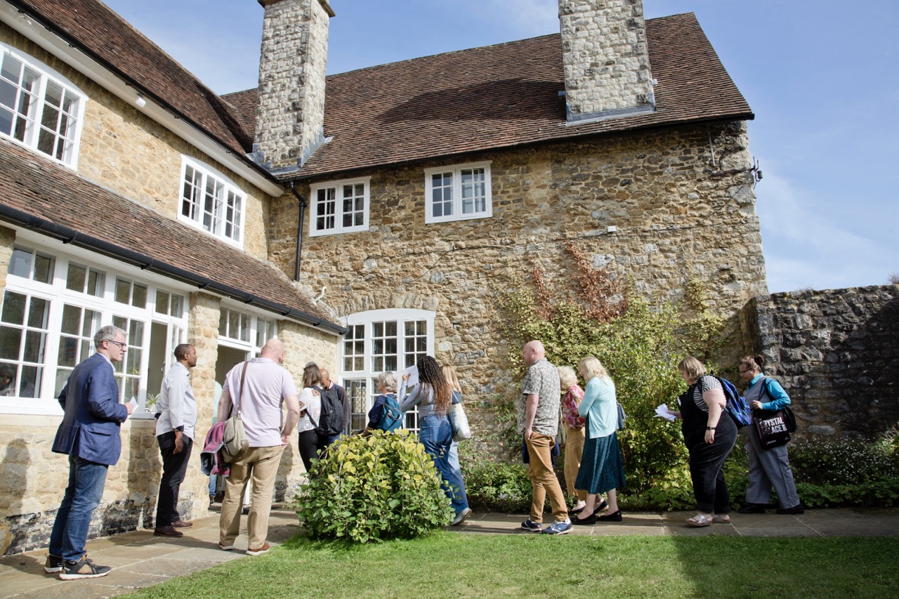 West Malling induction 2025 Around eight people, men and women walking and standing. They are standing on a path next to grass and an old brick building behind them. It's sunny.