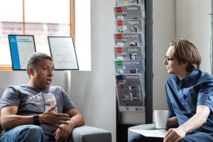 Two men chatting. They are indoors, sitting in armchairs in a waiting room or office.
