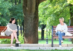 Two women smiling at each other in a park. They are sitting next to each other but on separate benches. It's a sunny day.