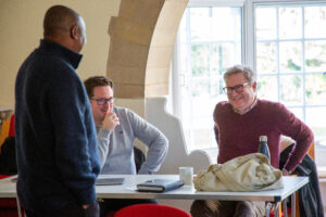 Three men smiling in a room. Two are sitting down, the other standing up.